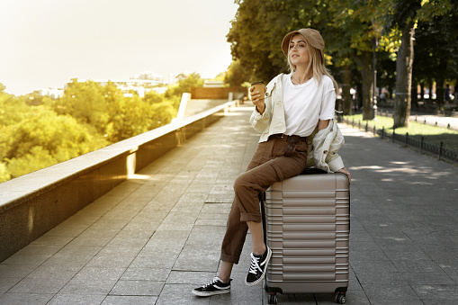 tourist woman in a European city, tourism in Europe. young woman sits on a large suitcase in the park with coffee in her hands. Disposable paper cup. Copy space.