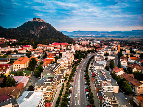 Color image depicting a high angle drone view of the city of Deva, a town in the Transylvania region of Romania. We can see communist-style apartment blocks and Deva's ancient citadel in the distance. The scene is set off by a sunset and cloudscape.