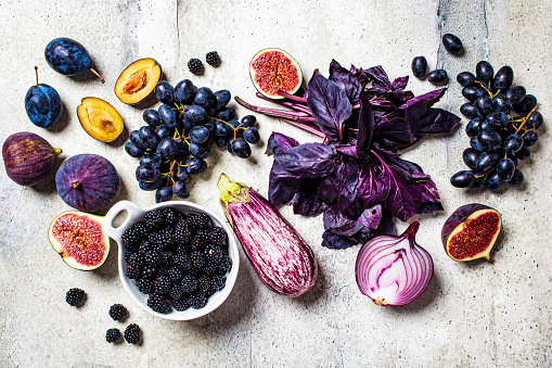 Raw purple vegetables and fruits on a gray concrete background. Flat lay purple food. Eggplant, grapes, figs, plums, blackberries, onions and basil, top view.