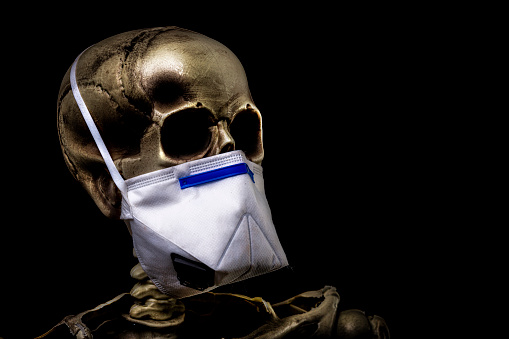 Human skeleton wearing a face mask skull in dramatic low key light, corona conid-19 against pitch black background