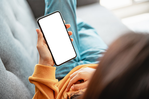Woman using mobile smartphone with blank white screen on a sofa in living room.