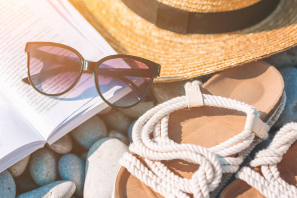 istock Beach hat on opened book with sunscreen and shoes on pebble beach 1272381226