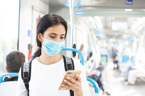 Coronavirus, COVID-19 spread prevention.
Student girl with protective face mask using smart phone and looking through the window while commuting by bus.