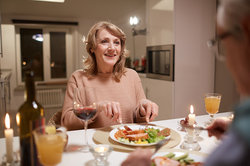 Happy mature woman sitting at the table eating dinner and smiling to her husband during their date