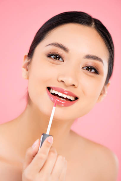 istock brunette asian woman looking at camera while applying lip gloss isolated on pink 1272392514