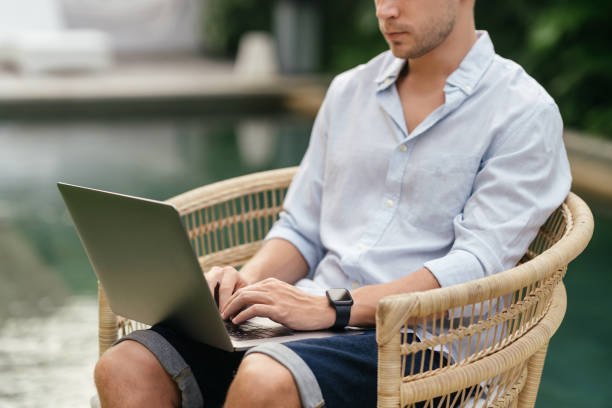 istock Young man using a laptop computer in a garden with a swimming pool. 1272394852