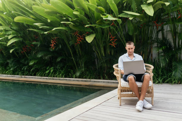 istock Young man using a laptop computer in a garden with a swimming pool. 1272394925
