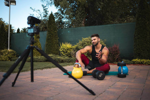 istock Male athlete sitting on exercise mat and making vlog at backyard. 1272395760