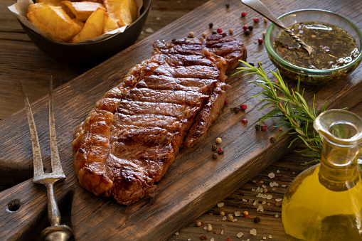 Grilled strip steak New York steak on cutting board over  rustic wooden board with sauce spices and french fries