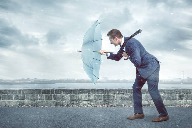 Businessman with an umbrella is facing strong headwind A young business man holding an umbrella to shield himself against a strong headwind. Conceptual image depicting adversity. endurance stock pictures, royalty-free photos & images