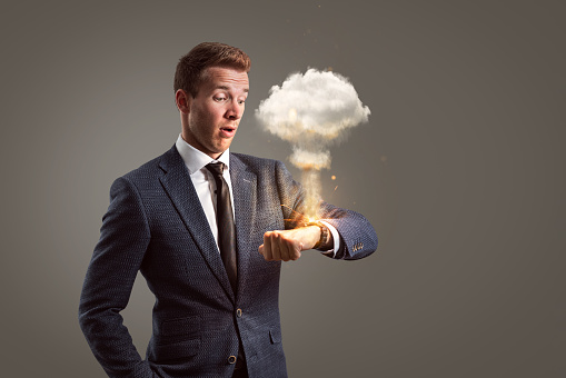 A young businessman is looking at his wrist watch. The watch is exploding while forming a large mushroom cloud. Conceptual image depicting time pressure or stress.