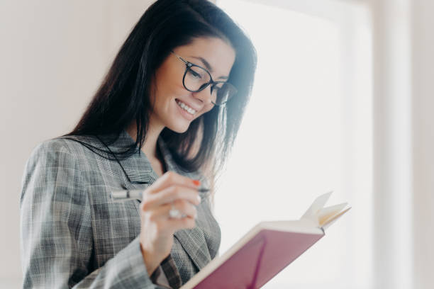 istock Sideways shot of pleasant looking brunette woman has happy expression makes notes in planner, does remote job, creats planning, wears optical eyeglasses and formal clothes, poses near window 1272404884