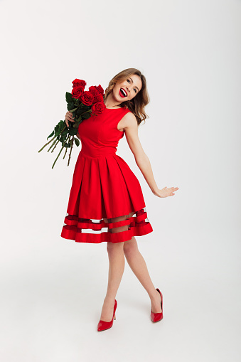 Full length portrait of a lovely young woman dressed in red dress holding bouquet of roses while standing and looking at camera isolated over white background
