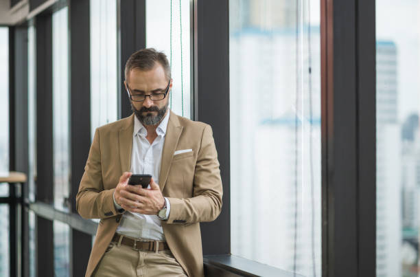 istock Young handsome business bearded man standing post near window using smartphone in office work place. 1272410161