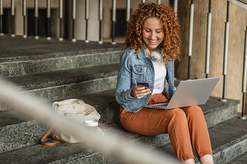 Young female student sitting on the stairs in front of university and using laptop and smart phone