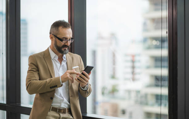 istock Young handsome business bearded man standing post near window using smartphone in office work place. 1272411014