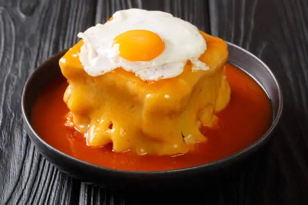 Photo of Portuguese delicious Francesinha sandwich with meat sausages, melted cheese, drizzled with tomato beer sauce and topped with a fried egg close-up. horizontal