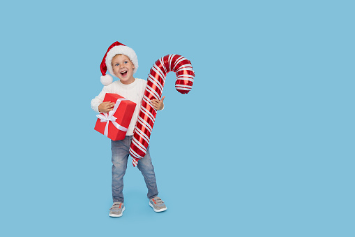 Portrait of a satisfied little child boy in christmas Santa hat. laughing isolated over blue background. Holds a gift box and big candy cane. Preparing for the New Year holidays