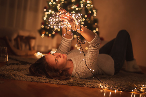 Young woman lying on the floor by nicely decorated Christmas tree relaxing at home and enjoying winter holoday season, holding bunch of Christmas lights