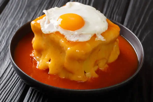 Photo of Hot Francesinha sandwich with sausages and meat, melted cheese, poured with tomato beer sauce and topped with a fried egg close-up in a plate. horizontal