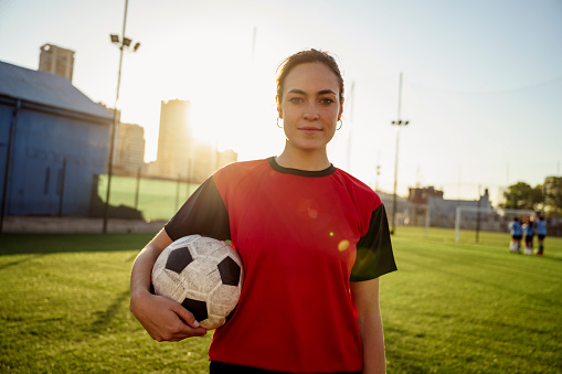 Lens flare close-up of young Buenos Aires female footballer in red and black uniform looking at camera with ball under her arm.
