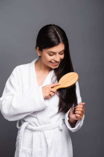 brunette woman in bathrobe brushing hair and smiling isolated on black brunette woman in bathrobe brushing hair and smiling isolated on black brushing hair stock pictures, royalty-free photos & images
