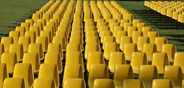 istock Long rows of empty yellow plastic chairs geometrically arranged on a lawn. 1272422690