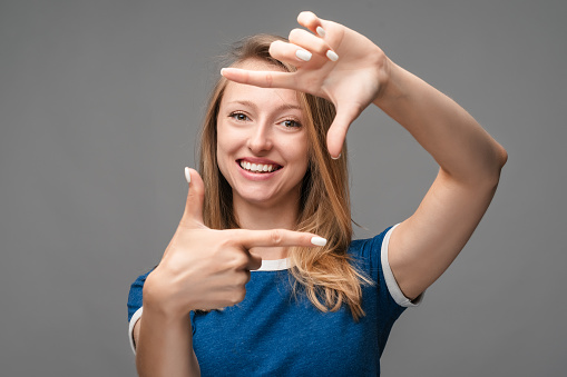 Photo of funny blonde woman in casual blue t shirt, smiling and making photo frame with fingers. Human emotions, facial expression concept. Studio shot, white background