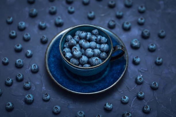 Fresh blueberries in a blue ceramic cup with a saucer on a dark background, a beautiful coffee cup filled with wild berries
