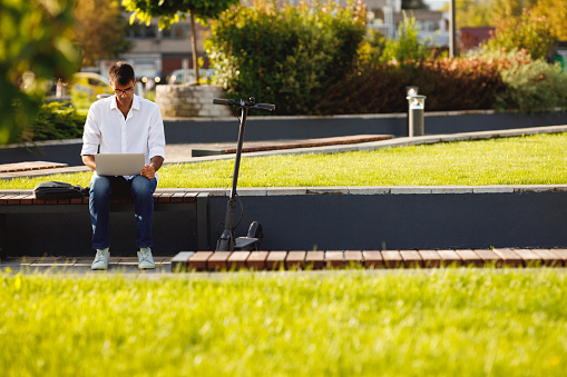 Young man in casual clothes sitting on a bench and using lap top with an electric scooter next to him.