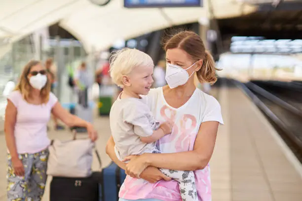 A young mother, wearing a protective mask or surgical mask, is waiting for the train at the train station with her son in her arms, photographed in color and in high resolution