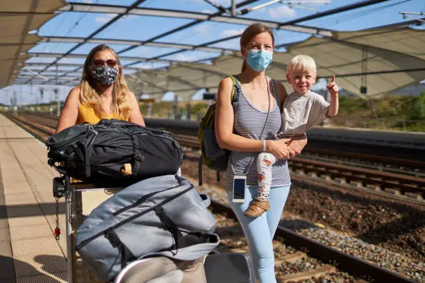 A young mother has her son in her arms and her mother pushes a baggage cart next to them on the platform. Both women are wearing protective masks or surgical masks, photographed in high resolution with copy space