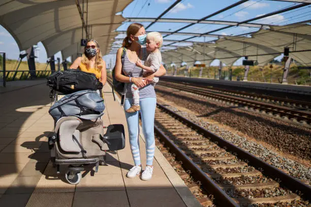 A young mother has her son in her arms and her mother pushes a baggage cart next to them on the platform. Both women are wearing protective masks or surgical masks, photographed in high resolution with copy space