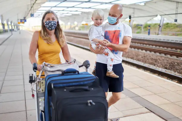 A senior woman with long hair and a facemask or surgical mask walks with her son and grandson at the station with their suitcases along the tracks to the next train, photographed in high resolution in color with copy space