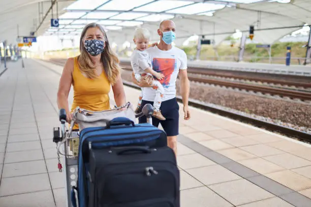 A young father is holding his son in his arms and his mother is pushing a luggage cart next to them on the platform. Both adults wear protective masks or surgical masks, photographed in high resolution with copy space