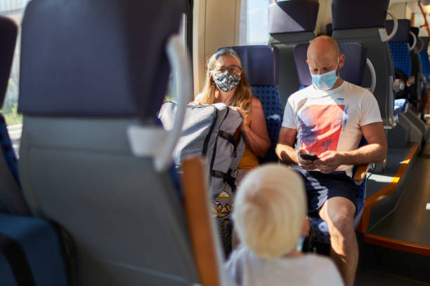 istock A middle-aged man looks at his smartphone or cell phone while wearing a protective mask or surgical mask and sits on a train next to an elderly woman who has her suitcase on her lap 1272435123