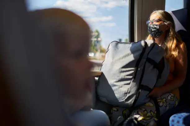 A senior citizen sits in a train with a large suitcase on her lap and a protective mask or surgical mask and looks out the window. Photographed in high resolution with copy space
