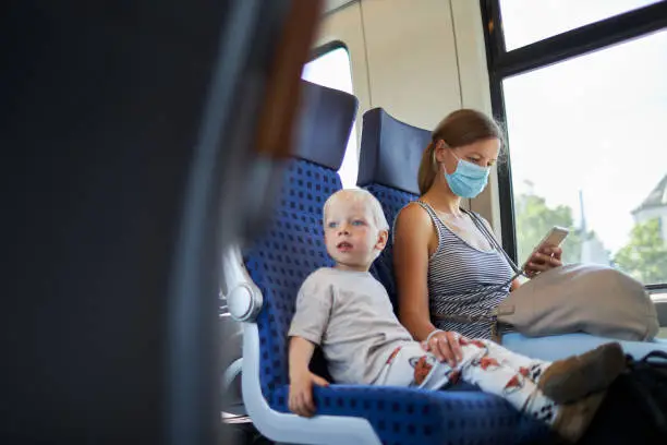 A blond little boy sits next to his mother, who is wearing a protective mask or surgical mask, on the train and looks to the left, photographed with copy space and in high resolution
