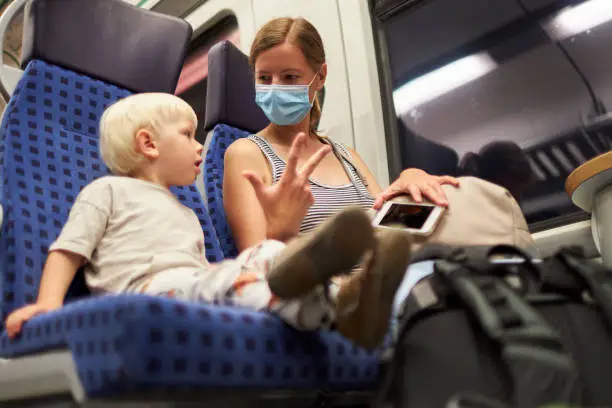 A blond little boy sits next to his mother who is wearing a protective mask or surgical mask and shows him with her fingers that the train will arrive in 3 minutes, on the train. photographed with copy space and in high resolution