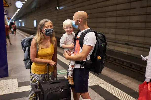 A senior woman with long hair and a facemask or surgical mask is waiting with her suitcase for the train together with her son and grandson at the train station, photographed in high resolution in color with copy space