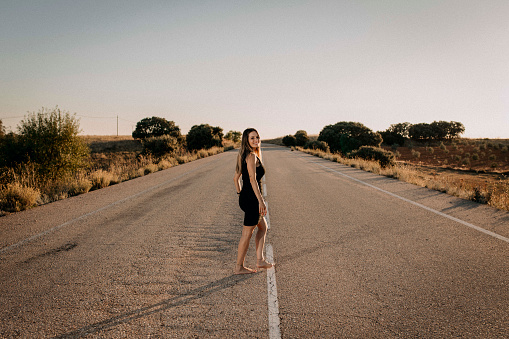 A photo of a smiling, barefoot woman in a black dress crossing an empty road.