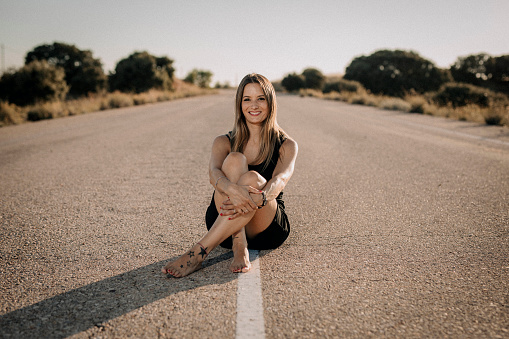 A photo of a smiling woman sitting, in a black dress, on an empty road.