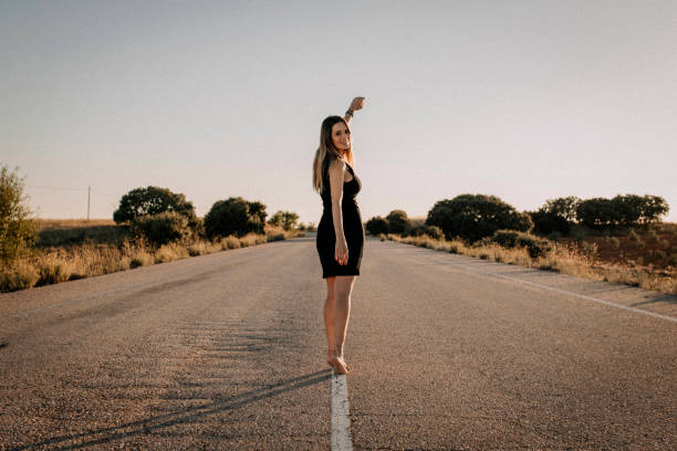 A photo of a smiling, barefoot woman in a black dress crossing an empty road.