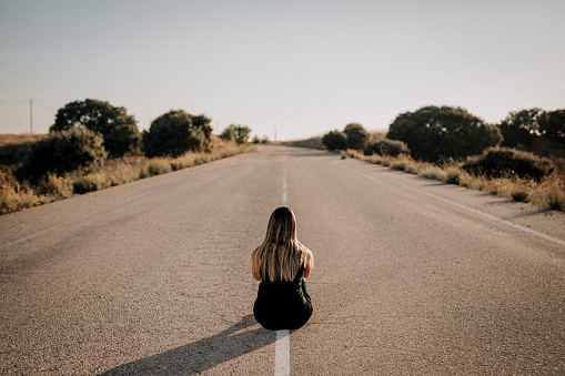 A photo of a woman sitting, in a black dress, with her back on an empty road.