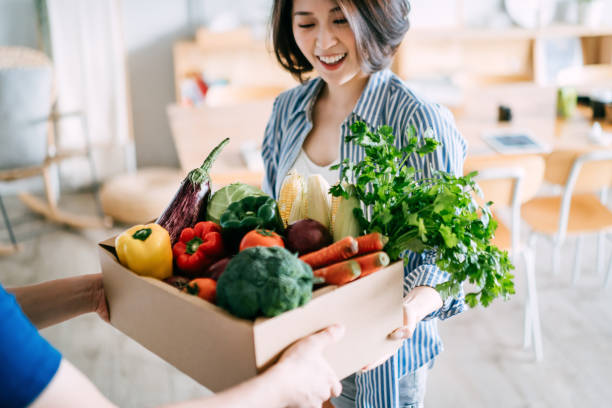 joven mujer asiática alegre que recibe una caja llena de alimentos orgánicos coloridos y frescos ordenados en línea de la persona de entrega en casa - veganismo fotos fotografías e imágenes de stock