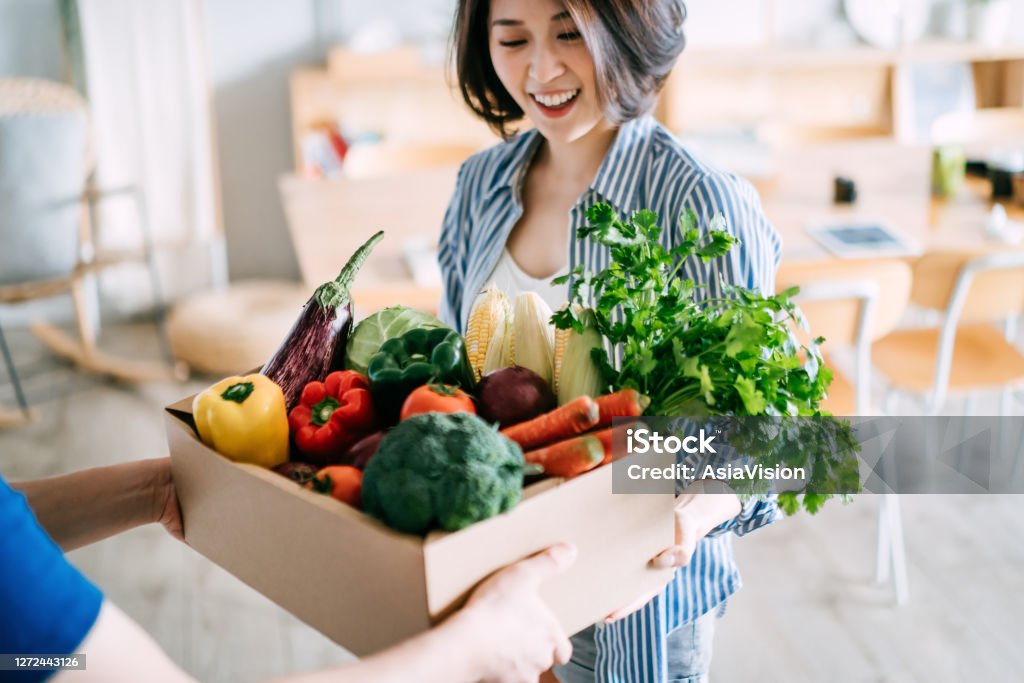 Cheerful young Asian woman receiving a box full of colourful and fresh organic groceries ordered online from delivery person at home Delivering Stock Photo