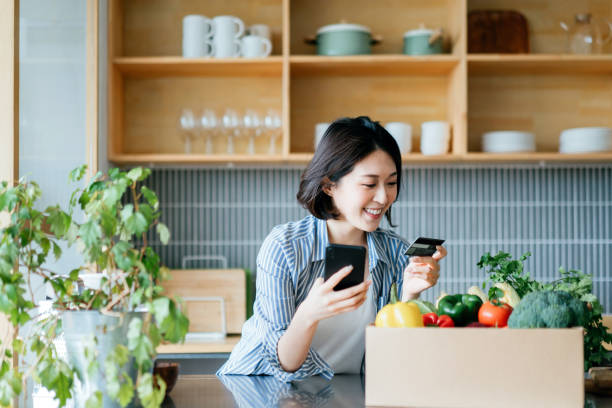 belle jeune femme asiatique souriante faisant ses courses en ligne avec l’appareil d’application mobile sur le smartphone et faisant le paiement en ligne avec sa carte de crédit, avec une boîte d’épicerie organique colorée et fraîche sur le comp - livraison à domicile photos et images de collection