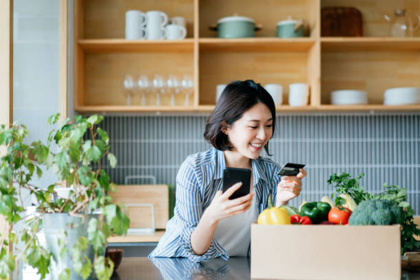 Beautiful smiling young Asian woman grocery shopping online with mobile app device on smartphone and making online payment with her credit card, with a box of colourful and fresh organic groceries on the kitchen counter at home Beautiful smiling young Asian woman grocery shopping online with mobile app device on smartphone and making online payment with her credit card, with a box of colourful and fresh organic groceries on the kitchen counter at home mobile payment photos stock pictures, royalty-free photos & images