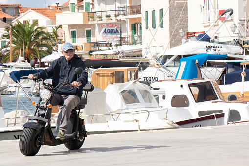 Vodice, Croatia - September 1, 2020: Senior man riding a wide wheel electric scooter with a seat, on the dock of  a small Mediterranean town