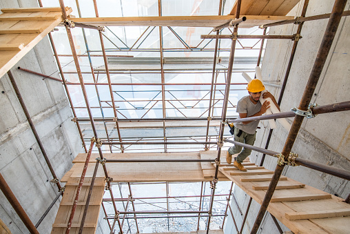 Photo of an African-American construction worker carrying plastic tubes while walking on scaffolding on a construction site.